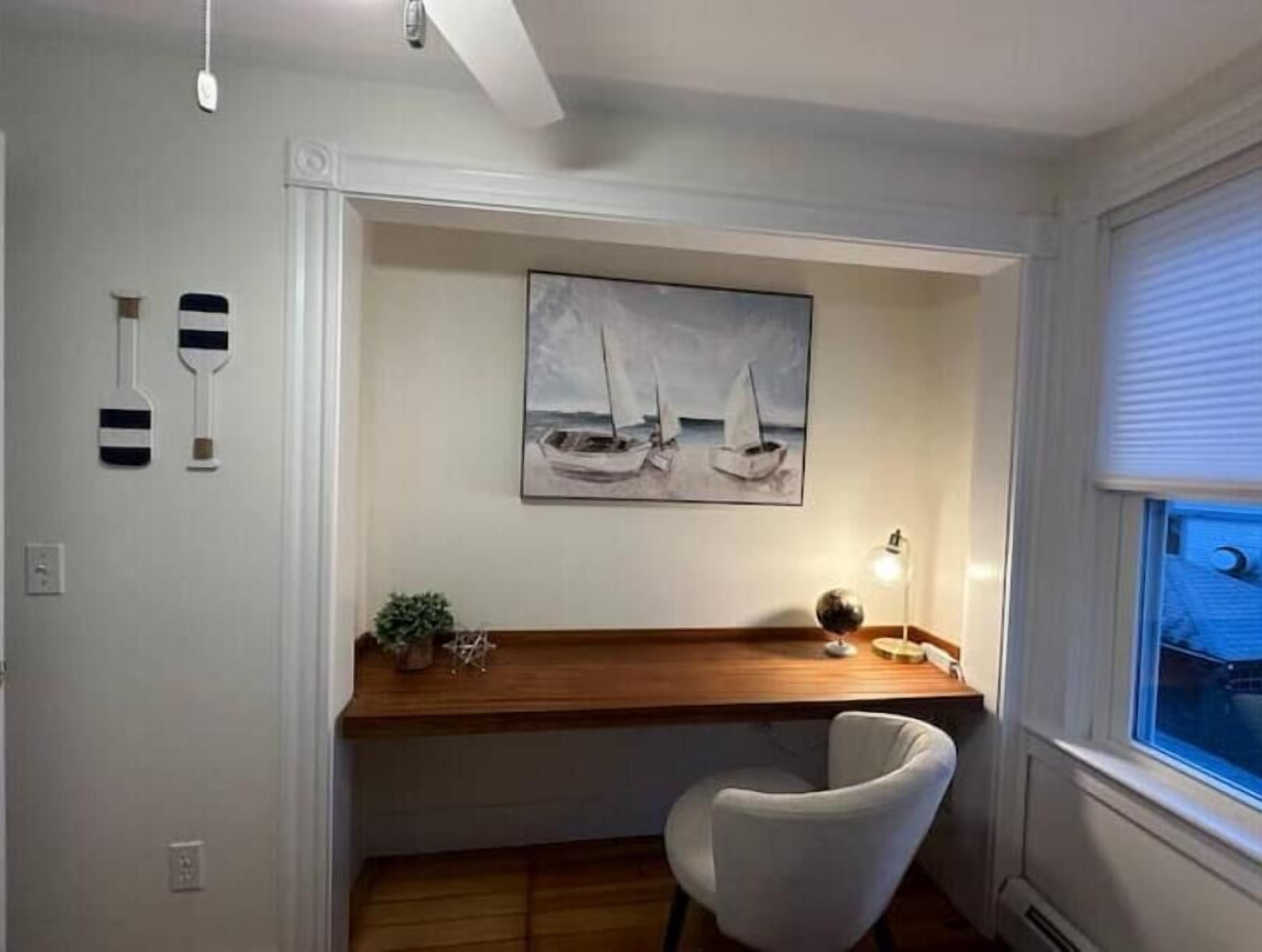 A compact room with a wooden desk, a white chair, a painting of sailboats, and a window with blinds.
