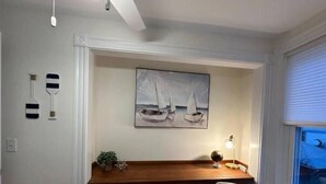 A compact room with a wooden desk, a white chair, a painting of sailboats, and a window with blinds. - Charming Cottage in Jamestown (Jamestown)