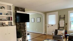 A living room with a stone accent wall, a flat-screen TV, a fireplace, and a cozy seating area with a coffee table. - Charming Cottage in Jamestown (Jamestown)