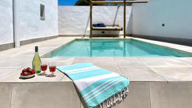 Une piscine avec un mur blanc et une terrasse en bois. Sur la terrasse, on peut voir une bouteille de vin, un verre et une assiette de fraises.