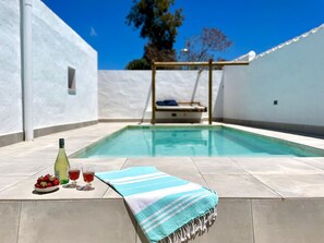 Una piscina con un muro blanco y una cubierta de madera. En la cubierta hay una botella de vino, una copa y un plato de fresas.