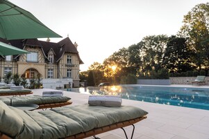 Uma área à beira da piscina com espreguiçadeiras e um grande guarda-sol, uma piscina e um edifício com estilo arquitetônico tradicional ao fundo.
