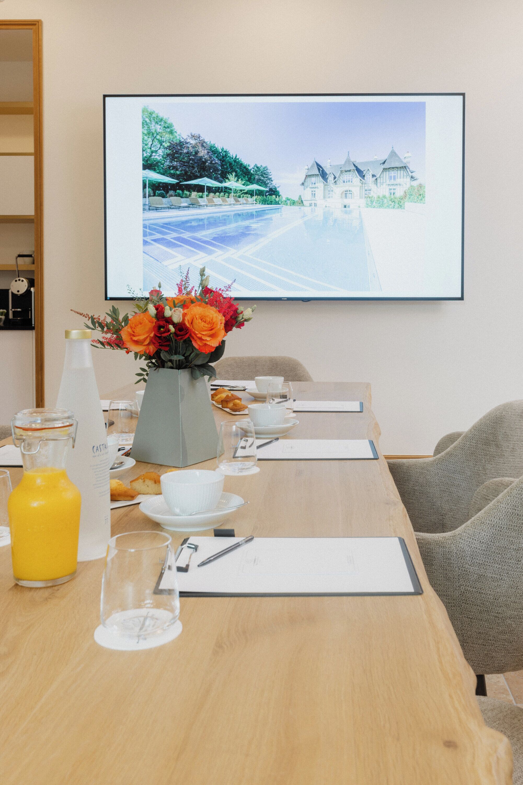 A conference room with a long table set for a meeting, a large screen displaying a scenic image, and a vase with flowers on the table.