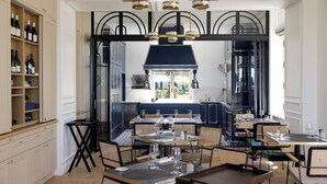 A restaurant with a central dining area, kitchen visible through an open archway, and shelves with bottles on the left. - Maison Douce Epoque - Deauville (Benerville-sur-Mer)