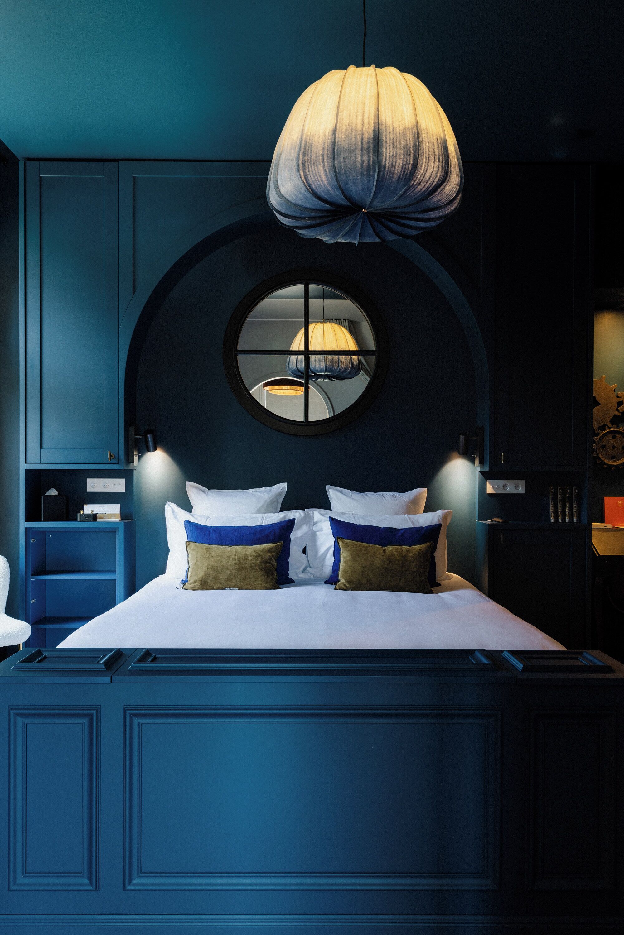 Une chambre avec un grand lit, un grand miroir rond au-dessus du lit et un luminaire suspendu.