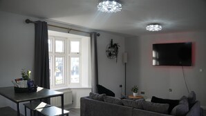 Un salon moderne avec un canapé gris, une table à manger avec deux chaises, un téléviseur fixé au mur et une fenêtre avec des rideaux.