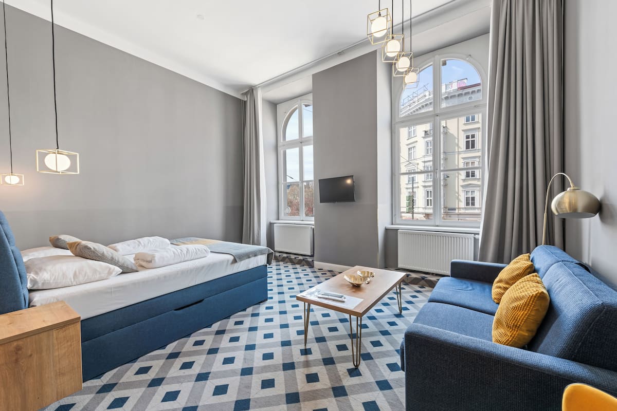 A modern hotel room with a blue sofa, a bed with white bedding, a wooden coffee table, and a large window with a view of buildings.