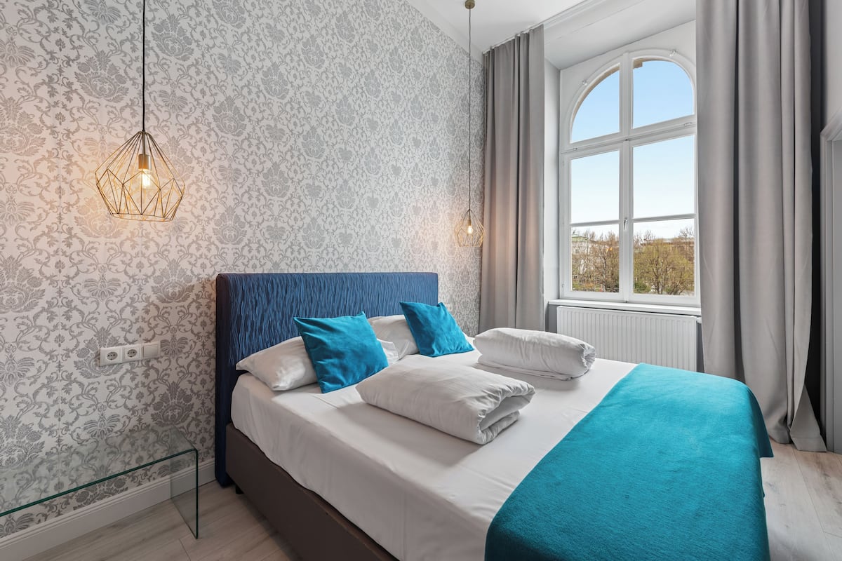 A neatly made bed with white and blue bedding, a geometric pendant light, and a window with curtains.
