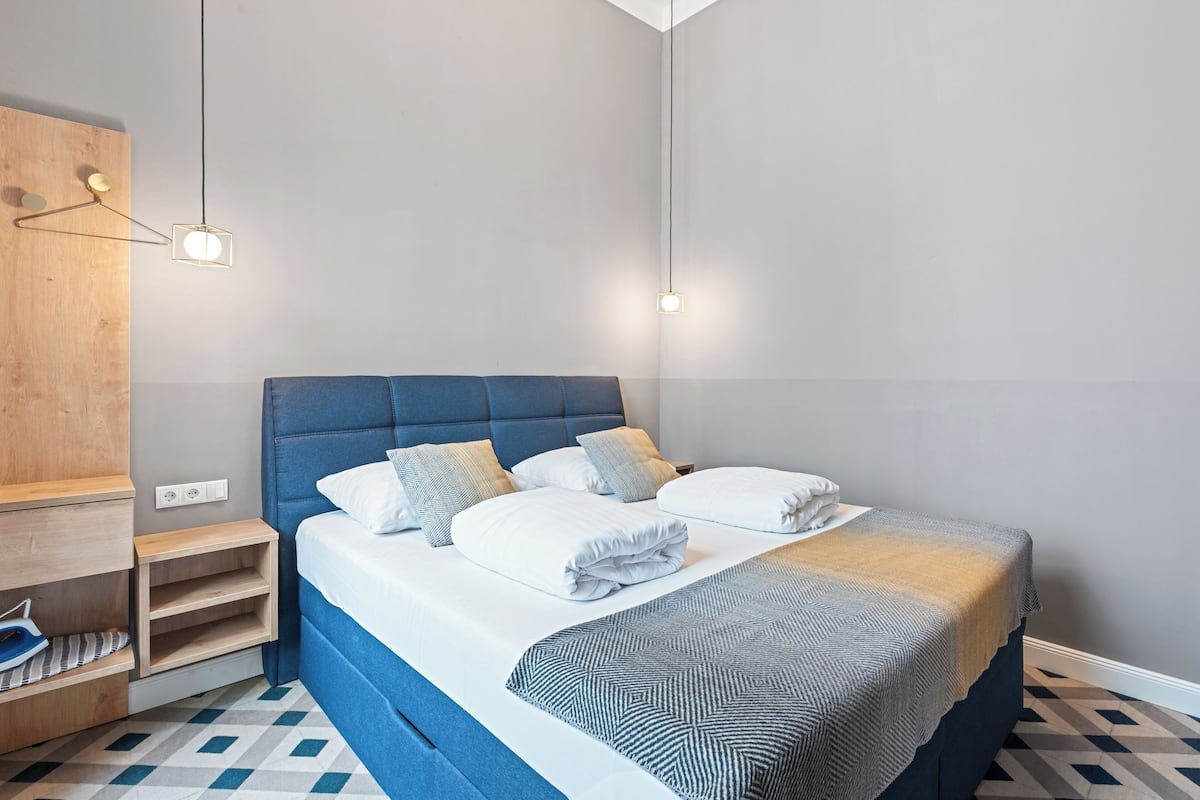 A modern bedroom with a blue headboard, a neatly made bed with white linens, and a geometric-patterned throw.
