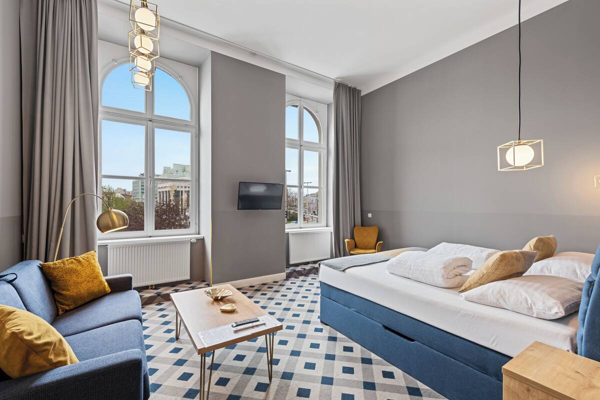 A hotel room with a bed, a sofa, a coffee table, a TV, and large windows.