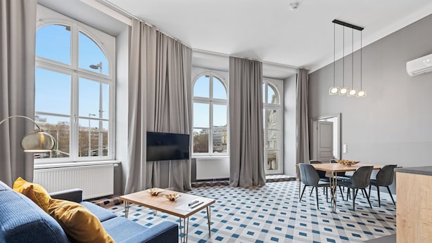 Een moderne woonkamer met een bank, televisie, eettafel en stoelen. Er zijn grote ramen met gordijnen, een kamer met een patroon en een plafondlamp.