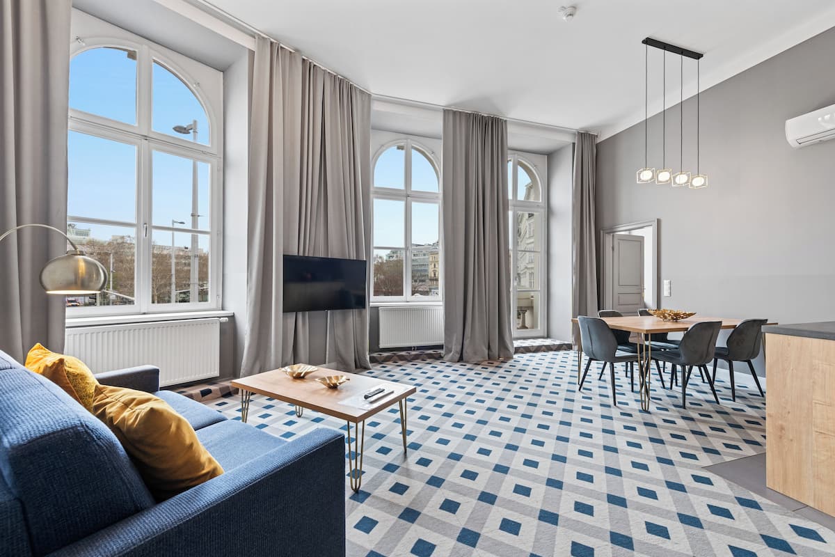 A modern living room with a sofa, TV, dining table, and chairs. There are large windows with curtains, a patterned floor, and a ceiling light fixture.