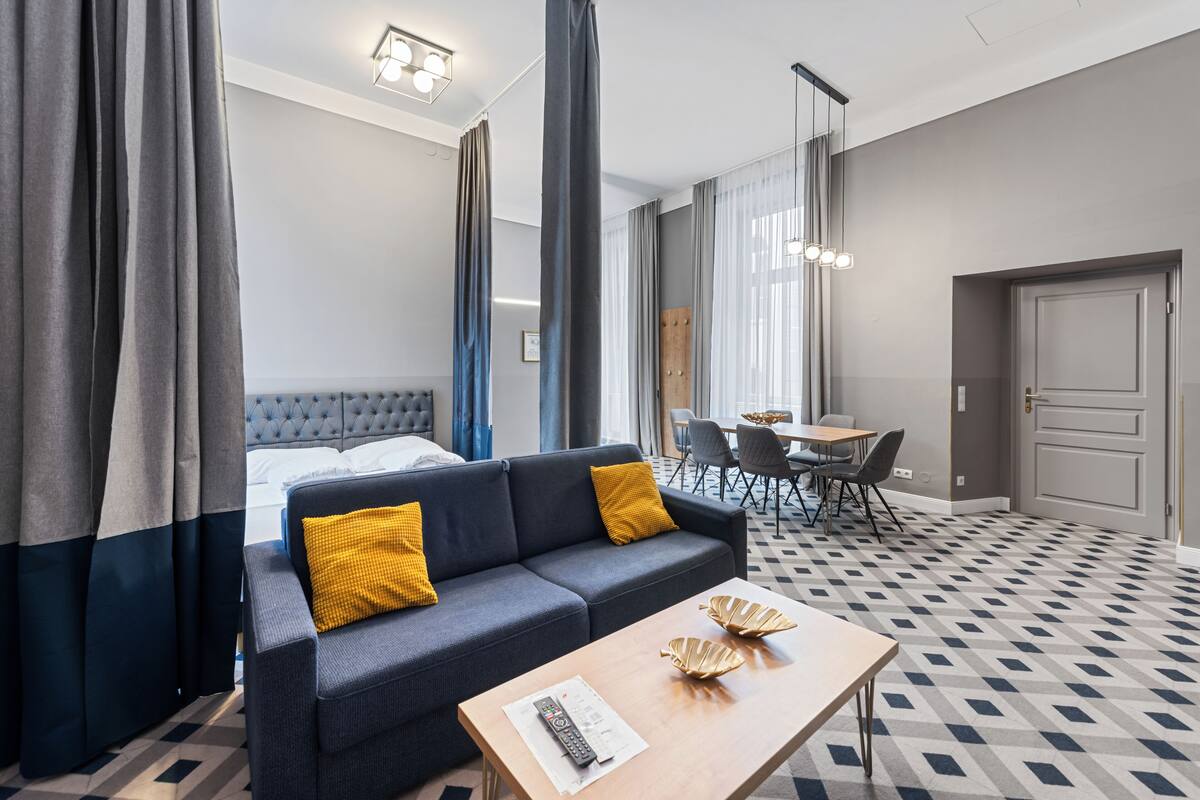 A hotel room with a grey sofa, a wooden coffee table, a dining area with a table and chairs, and a large window with curtains.