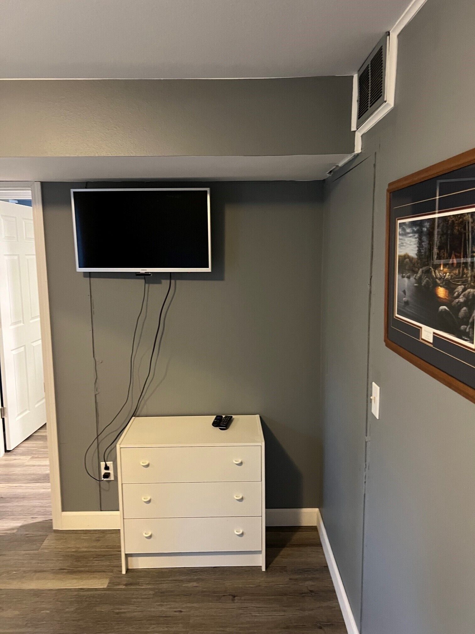 Ein Zimmer mit einem an der Wand montierten Fernseher, einer weißen Kommode und einem gerahmten Bild.