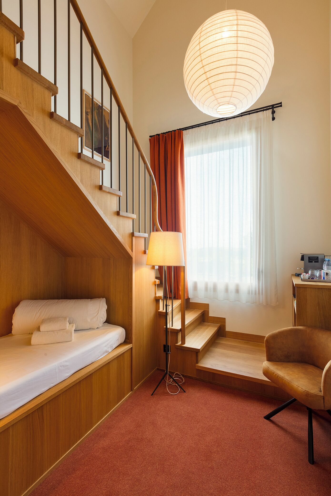 A hotel room with a wooden staircase, a bed with white linens, a standing lamp, a sofa, and a large window with curtains.
