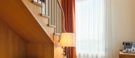 A hotel room with a wooden staircase, a bed with white linens, a standing lamp, a sofa, and a large window with curtains.