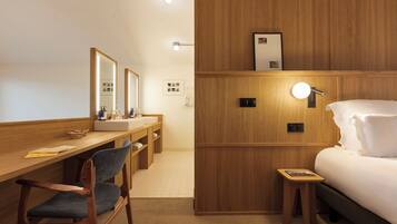 Quarto de hotel com escrivaninha de madeira, uma cadeira, cama, mesa de cabeceira, espelho e luminárias na parede.