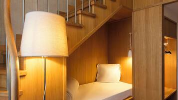 Habitación pequeña con una escalera de madera, una cama con colchón y almohadas blancos, una lámpara fijada a la pared y un estante pequeño.