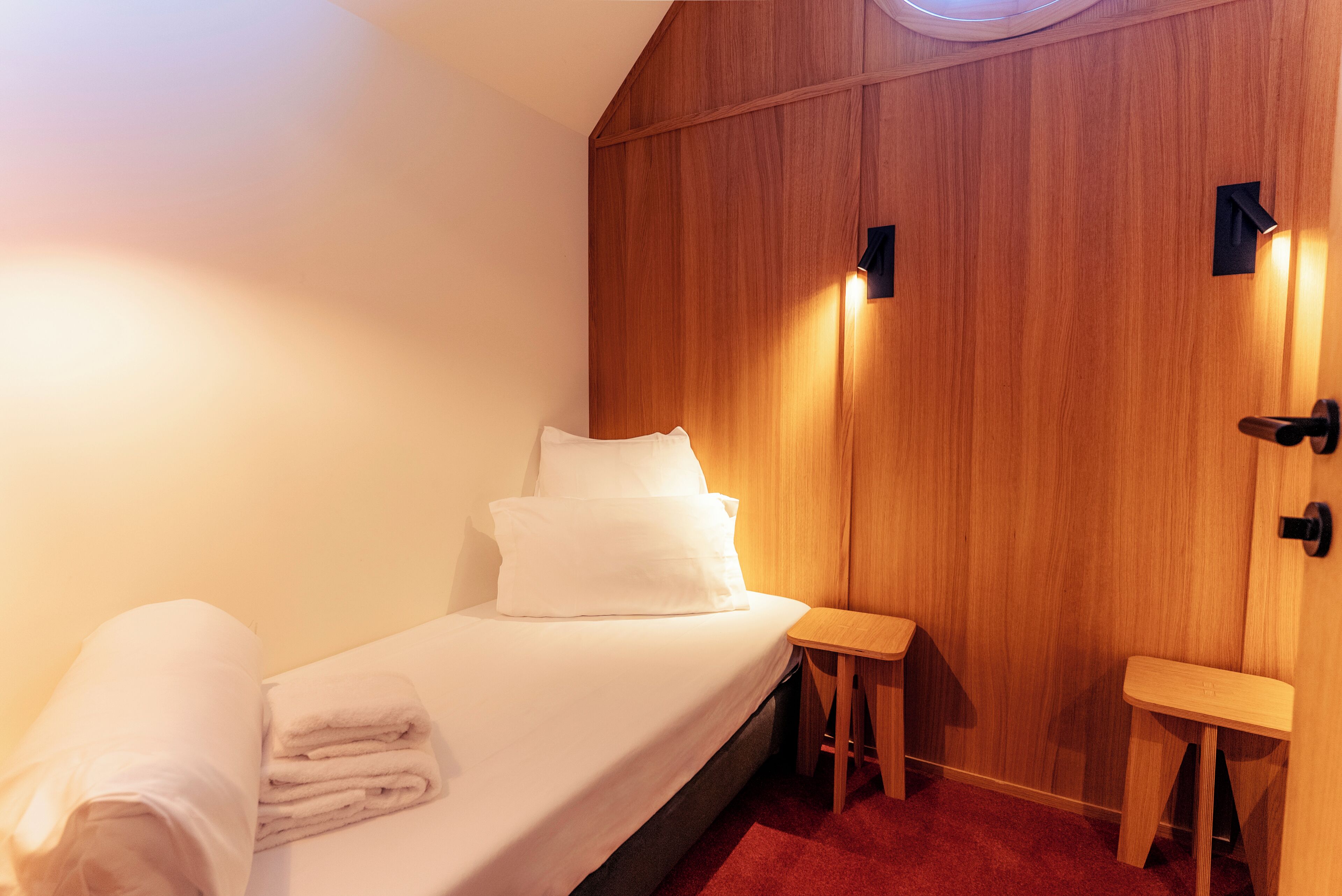 A single bed with white linens and pillows, a small wooden stool, and a wall-mounted light fixture.