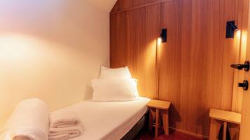 A single bed with white linens and pillows, a small wooden stool, and a wall-mounted light fixture.
