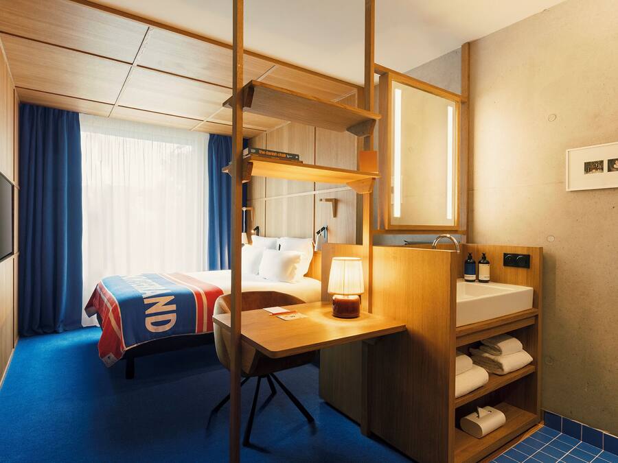 A hotel room with a bed, a desk, a mirror, a sink, and a shower area.