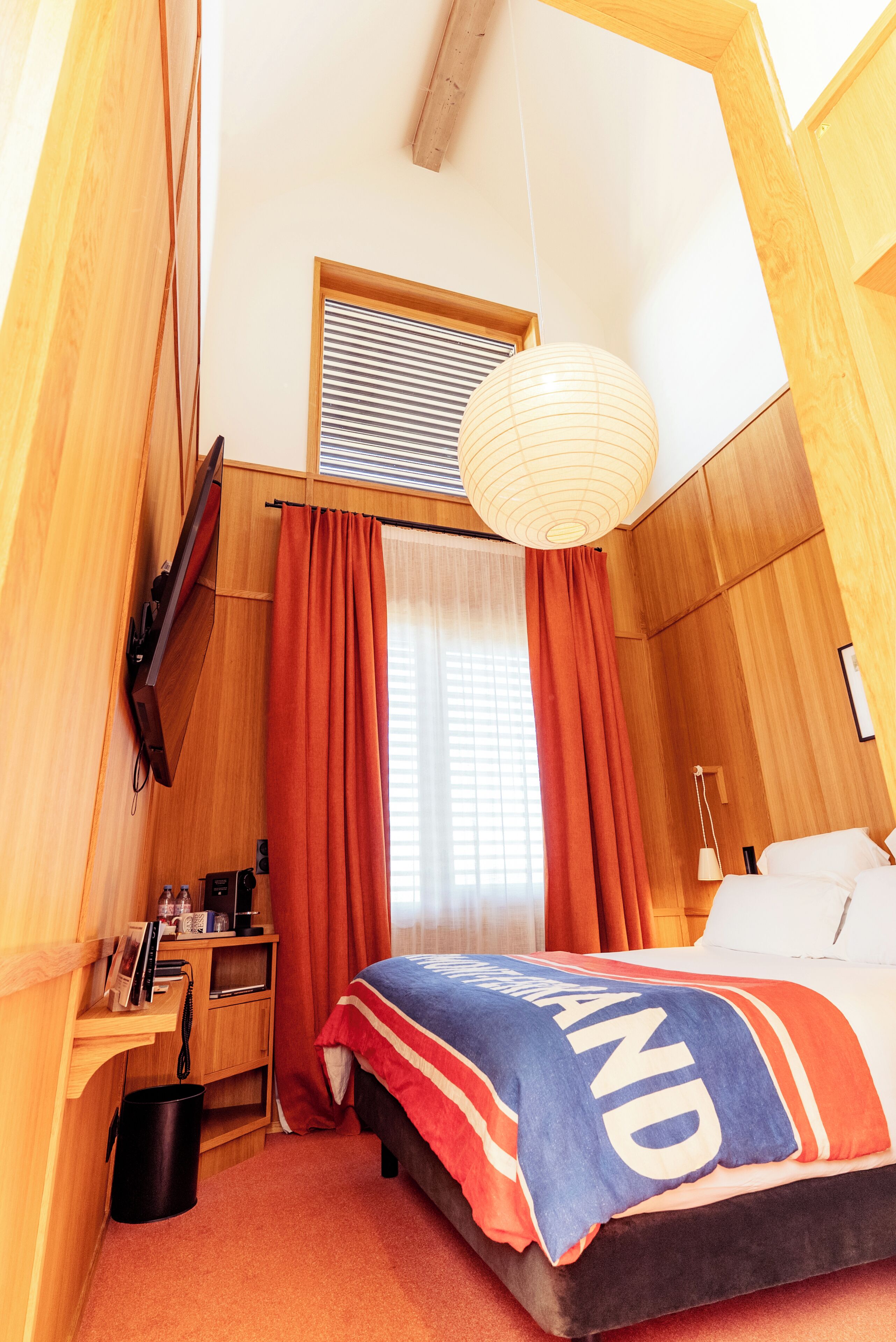 A bedroom with a bed, a desk, a hanging light fixture, and a window with curtains.