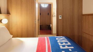 Un dormitorio con techo inclinado de madera, una cama con manta a rayas rojas y azules, y un baño visible al fondo.