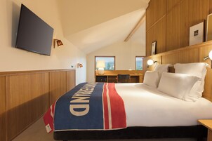 Habitación de hotel con cama, escritorio, silla, televisor y ventana con persianas.