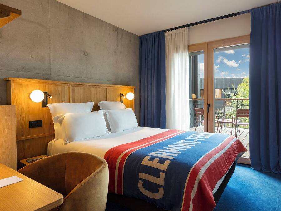 A hotel room with a bed, a wooden nightstand, a desk, and a chair. There is a balcony with a view of the sky and trees.