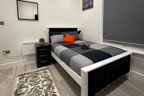 A modern bedroom with a bed, a nightstand, a small plant, and a window with a view. - Studio Flat in Ealing Broadway (London)