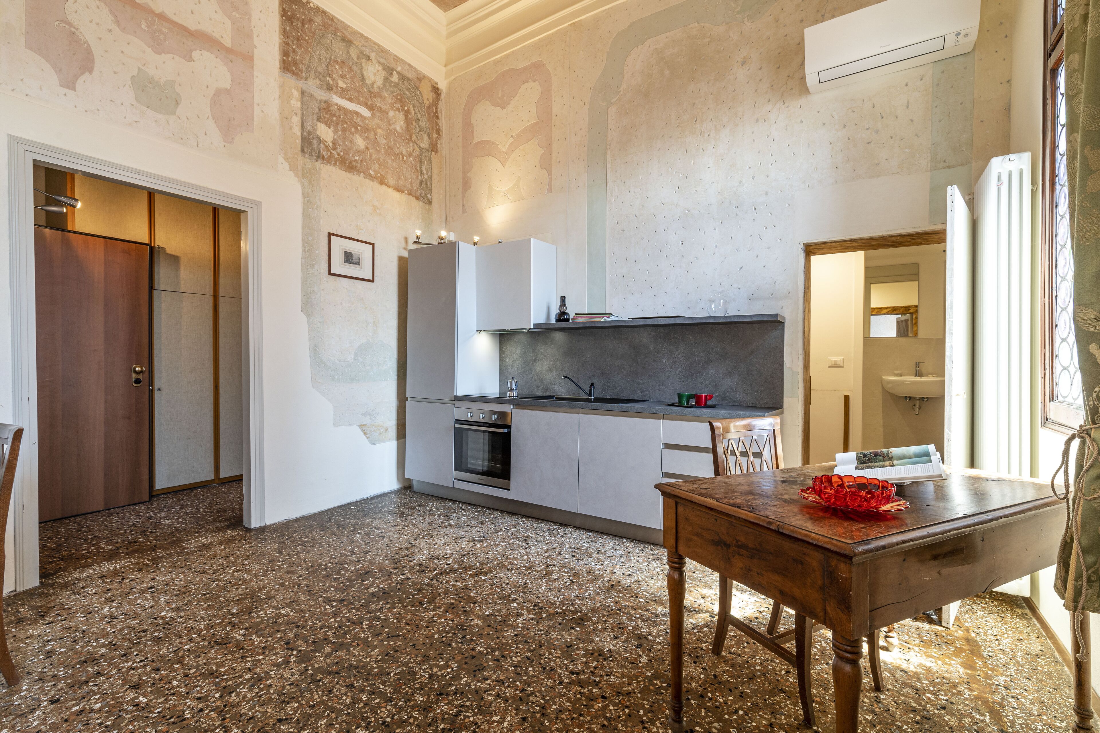 Una cocina compacta con horno empotrado, una mesa de comedor y un tazón rojo, y una puerta de madera.