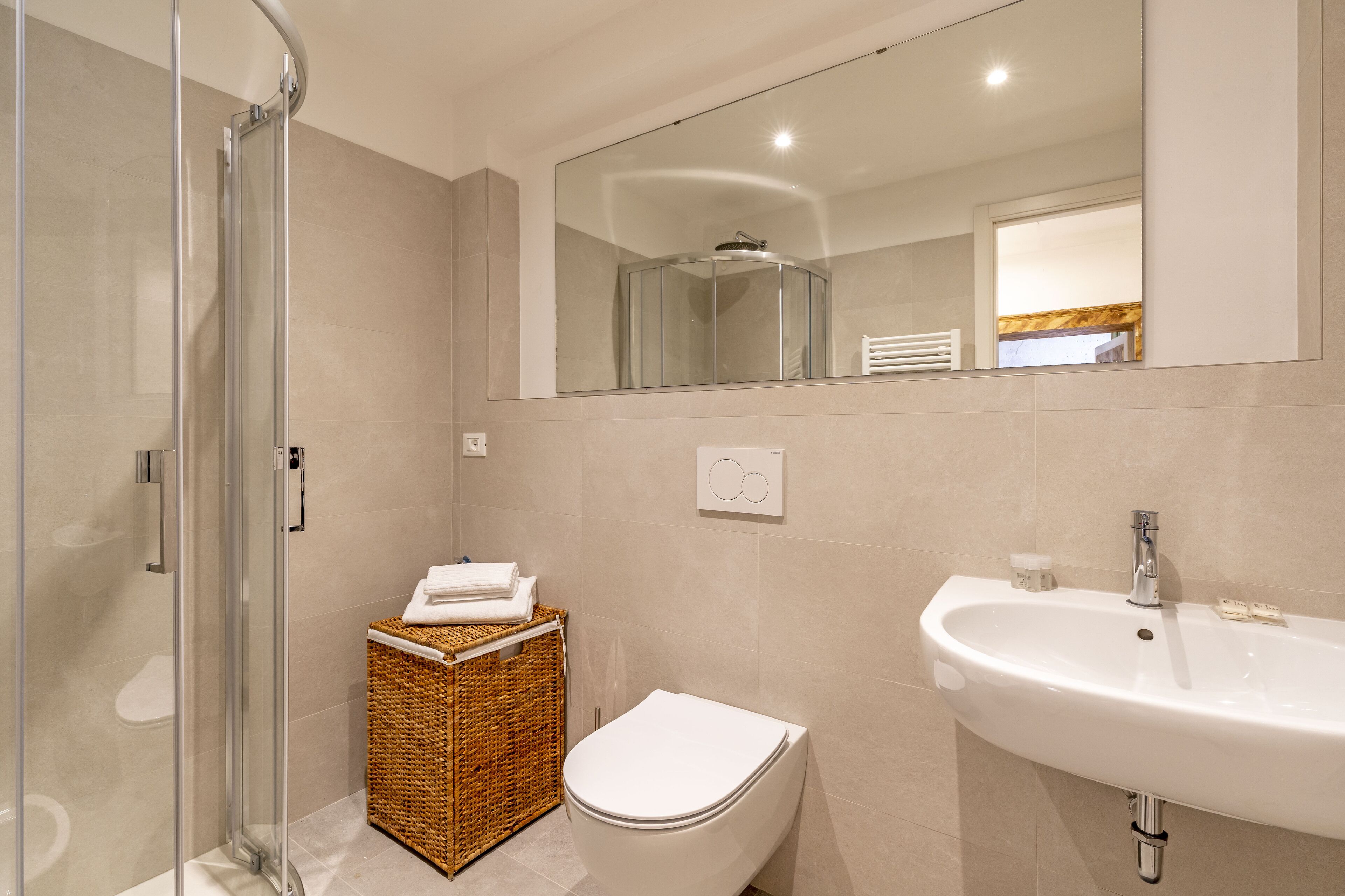 A modern bathroom with a glass shower enclosure, a white toilet, a wicker basket, and a large mirror above a sink.