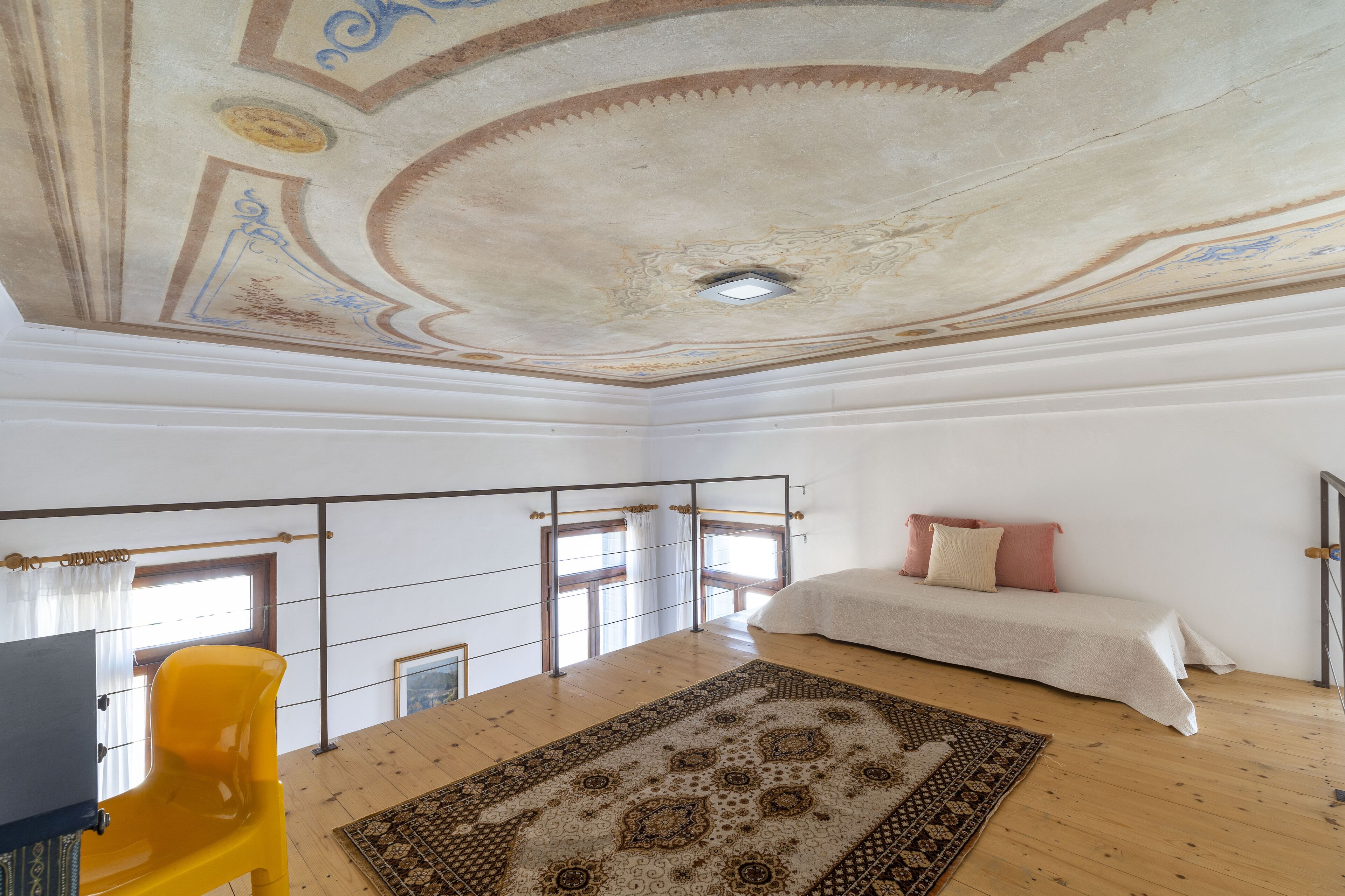 Un dormitorio con suelo de madera, alfombra estampada, cama con almohadas, una silla amarilla y techo con detalles decorativos.