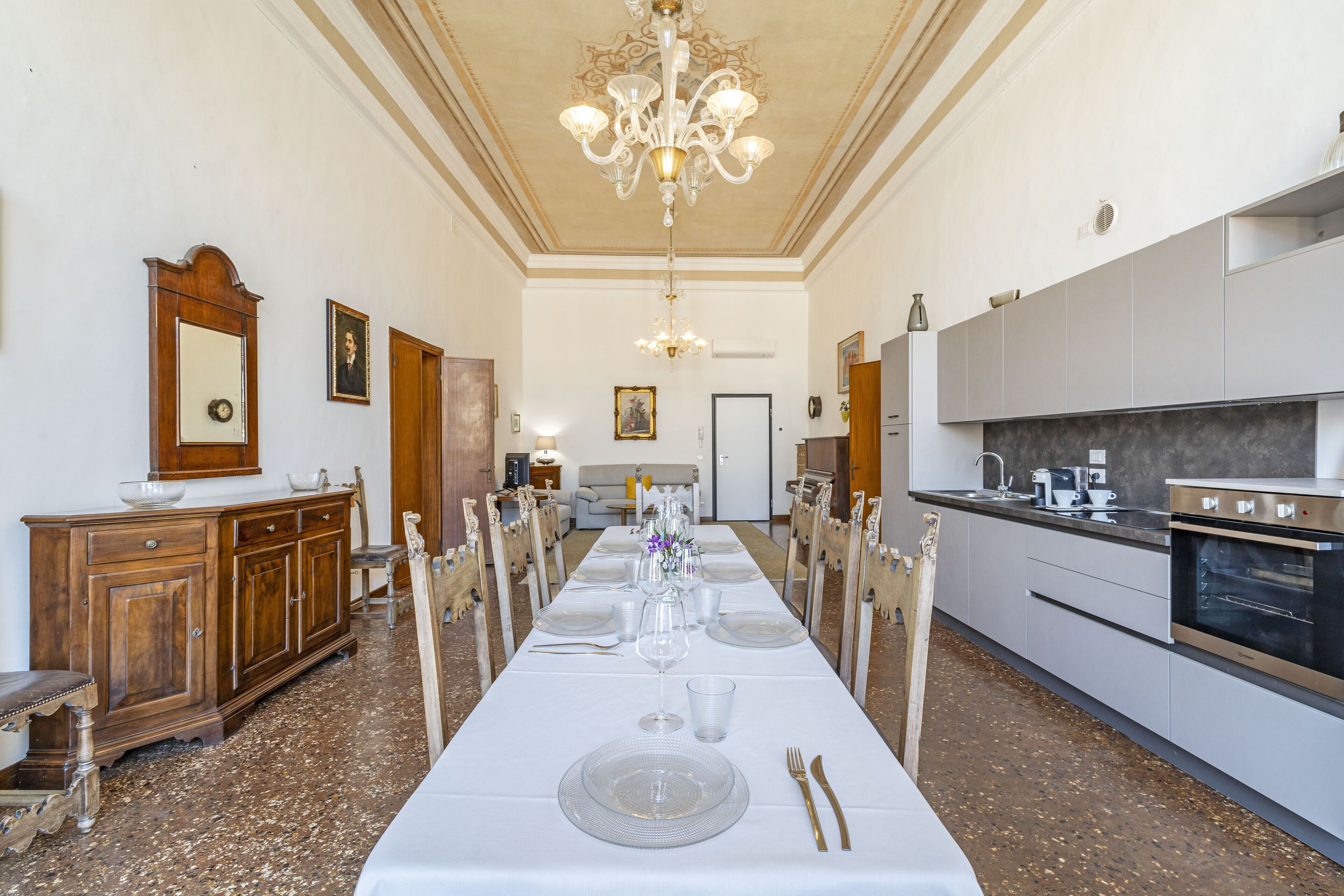 A long dining table set for a meal in a spacious room with a chandelier, wooden furniture, and a modern kitchen in the background.