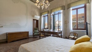 A room with a large bed, a wooden dresser, a desk, and two chairs. There are large windows with curtains, a chandelier, and a radiator. - Ca' Magia - Rialto Exclusive Residence by Wonderful Italy (Venice)