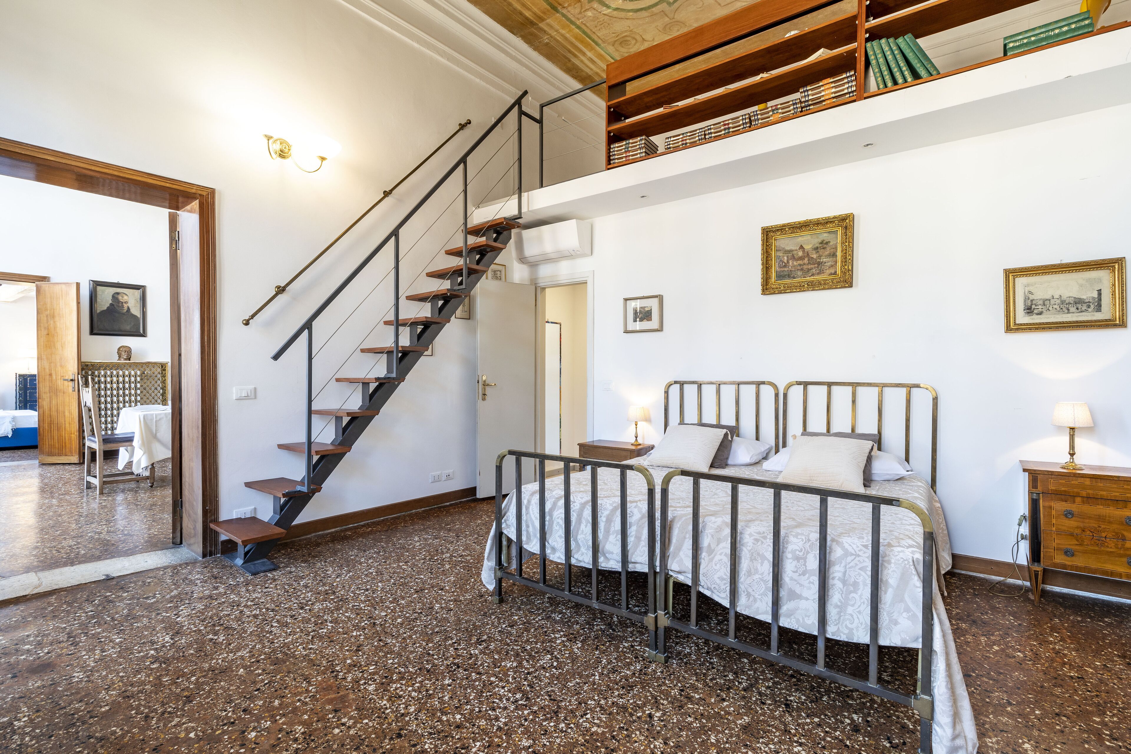 A bedroom with a bed, a nightstand, a lamp, a staircase, and a bookshelf.