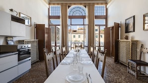 A dining room with a long table set for a meal, large windows, and a view of buildings outside. - Ca' Magia - Rialto Exclusive Residence by Wonderful Italy (Venice)