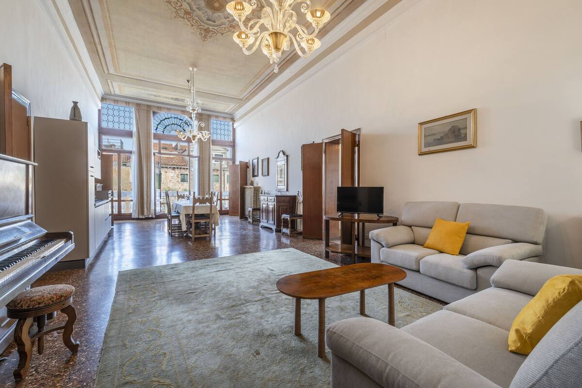 A spacious living room with a grand piano, a sofa set, a television, and a chandelier.