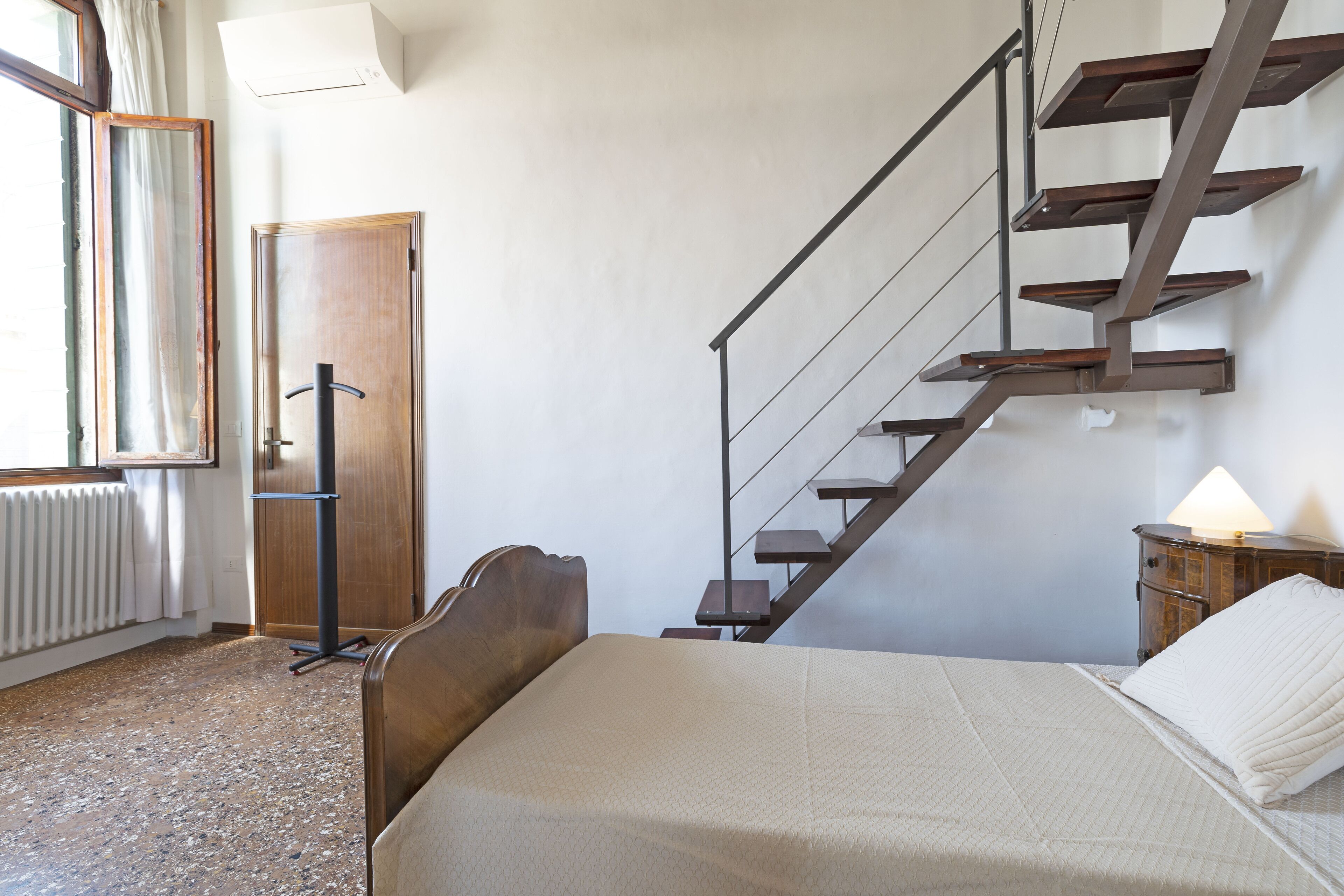 A bedroom with a bed, a wooden headboard, a window with curtains, a radiator, a wooden door, a staircase, and a nightstand with a lamp.
