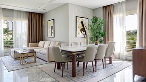 A modern living room with a dining area, featuring a marble floor, a beige sofa, a dining table with six chairs, and large windows with curtains.