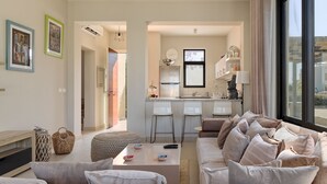 Een moderne woonkamer met een bank, voetenbankje en salontafel. Er is een keukenhoek met een koelkast, gootsteen en barkrukken. De kamer heeft grote ramen en uitzicht op de omgeving.