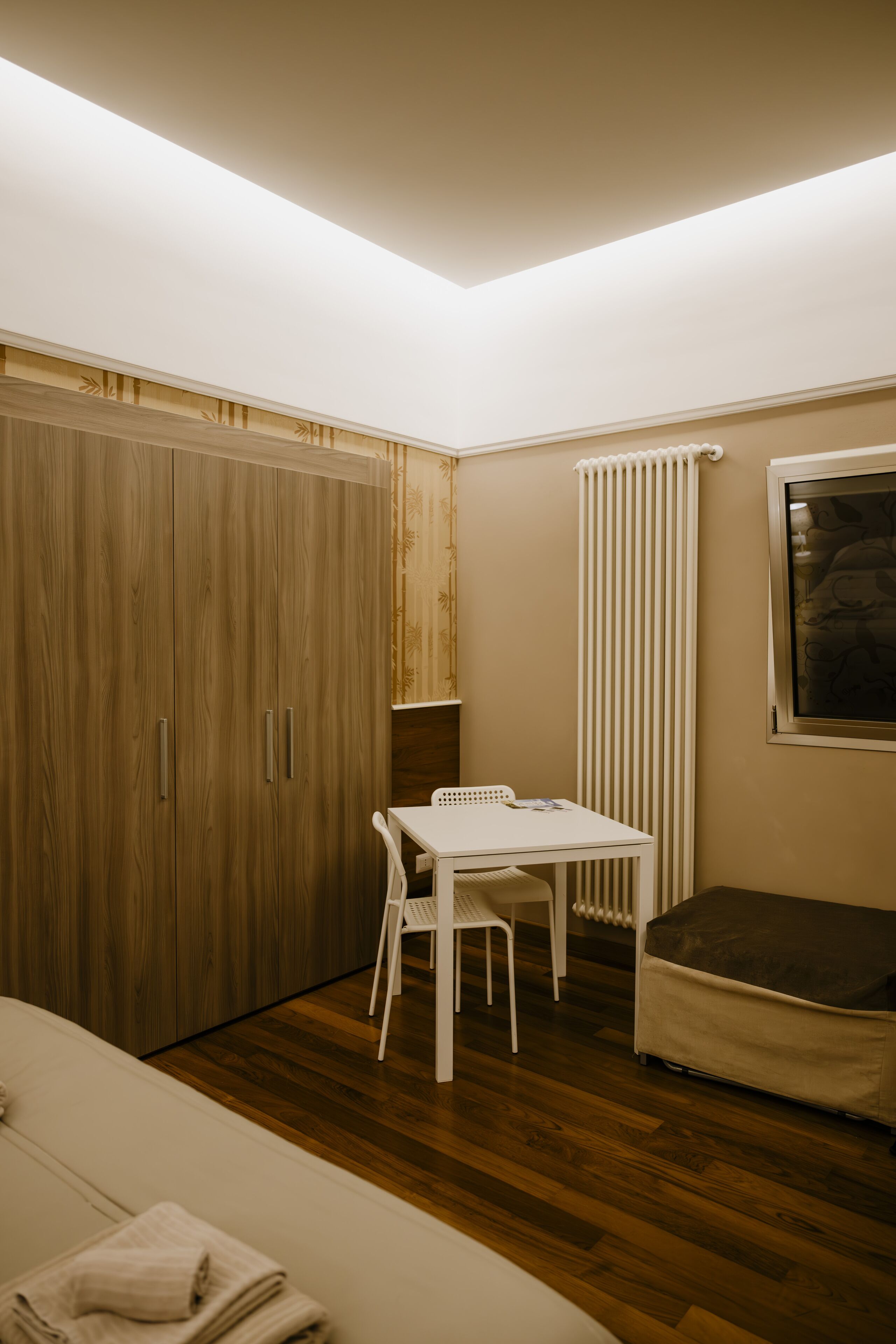 A room with a wooden wardrobe, a white table with chairs, a radiator, and a window.