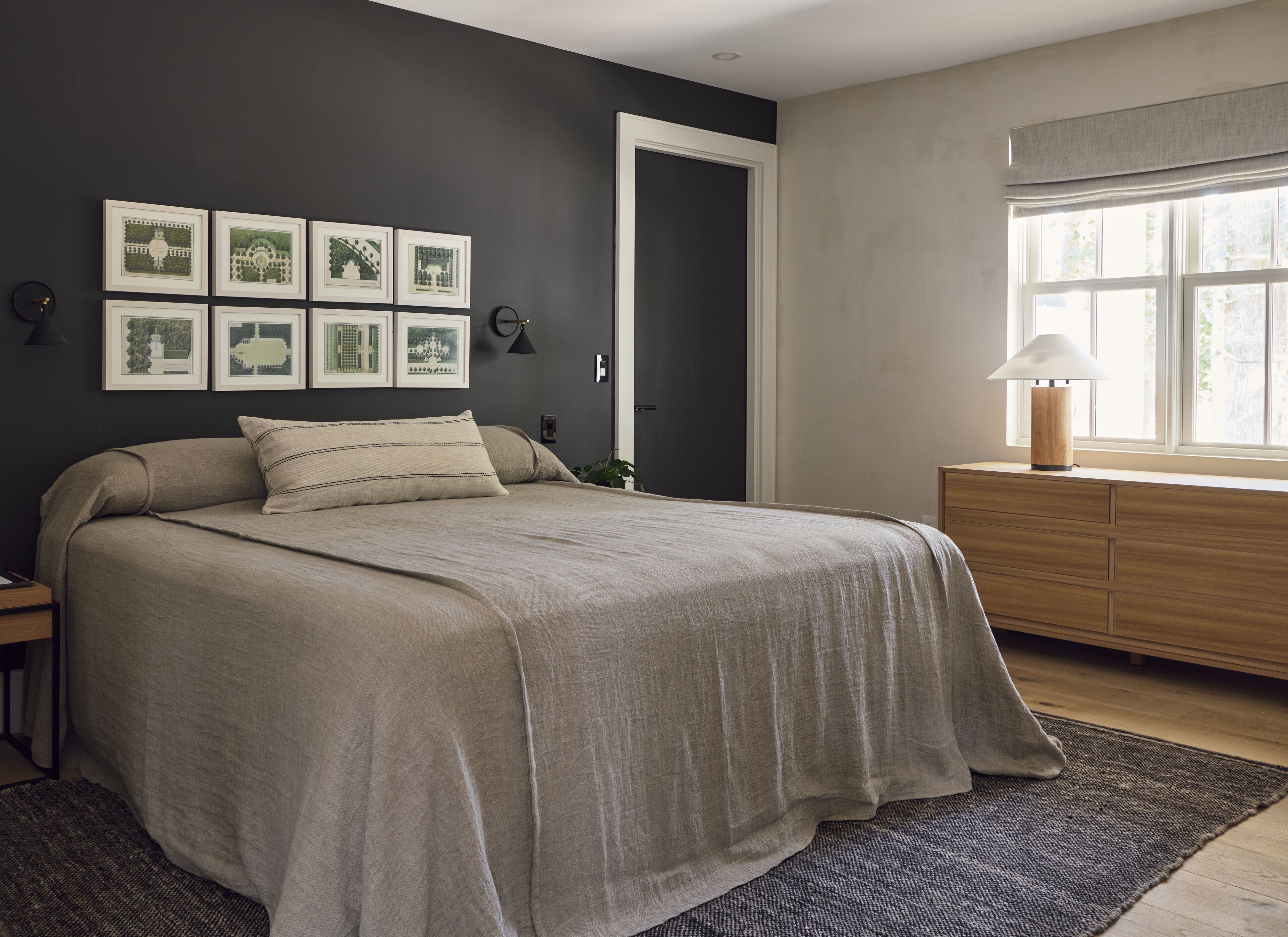 Un dormitorio con cama, tocador, mesita de noche, cuadros enmarcados en la pared y ventana con vistas.