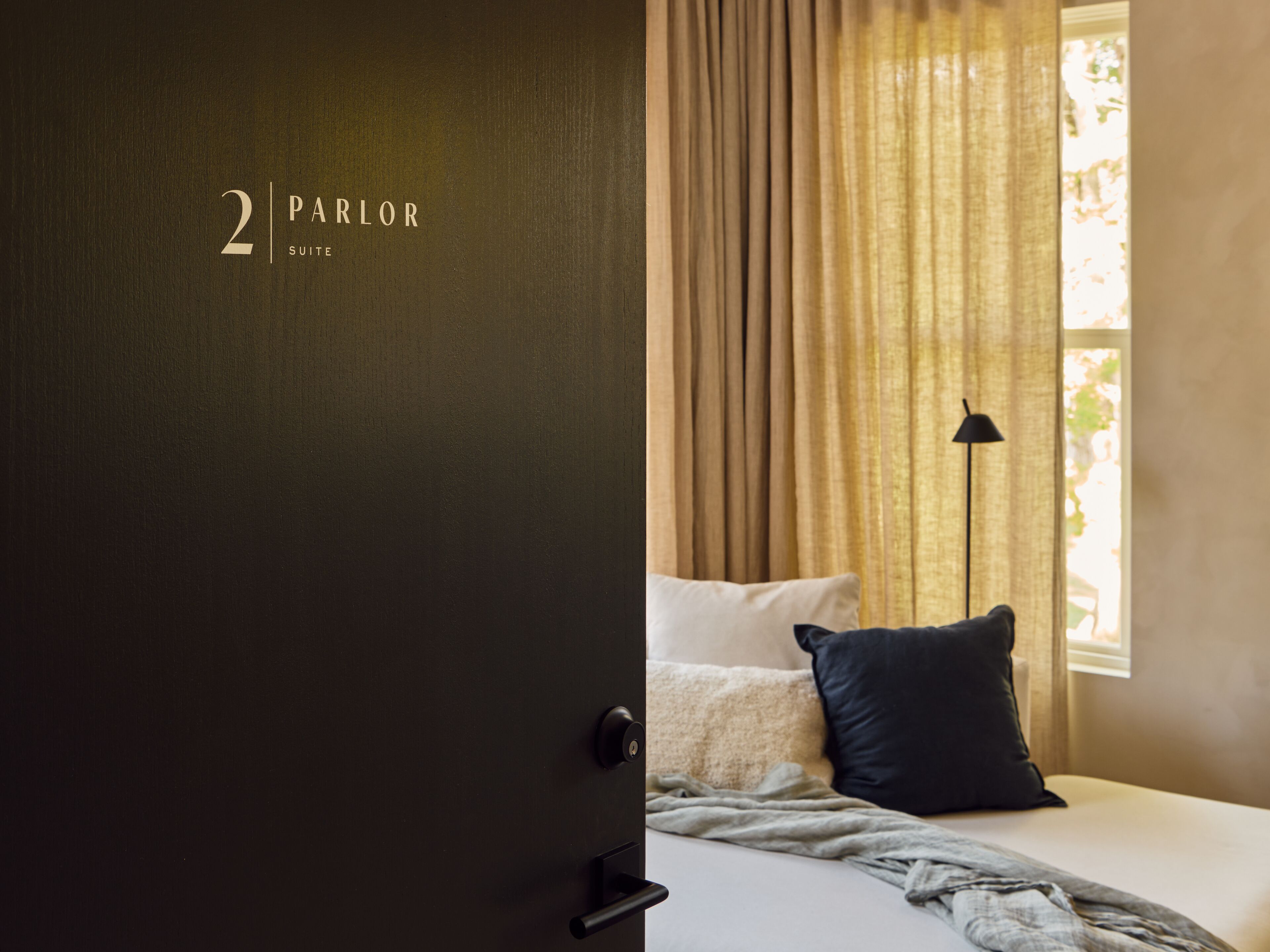 Une chambre d’hôtel avec une porte sombre, un lit avec une literie blanche, un oreiller foncé, un plaid gris et une fenêtre avec des rideaux beige.