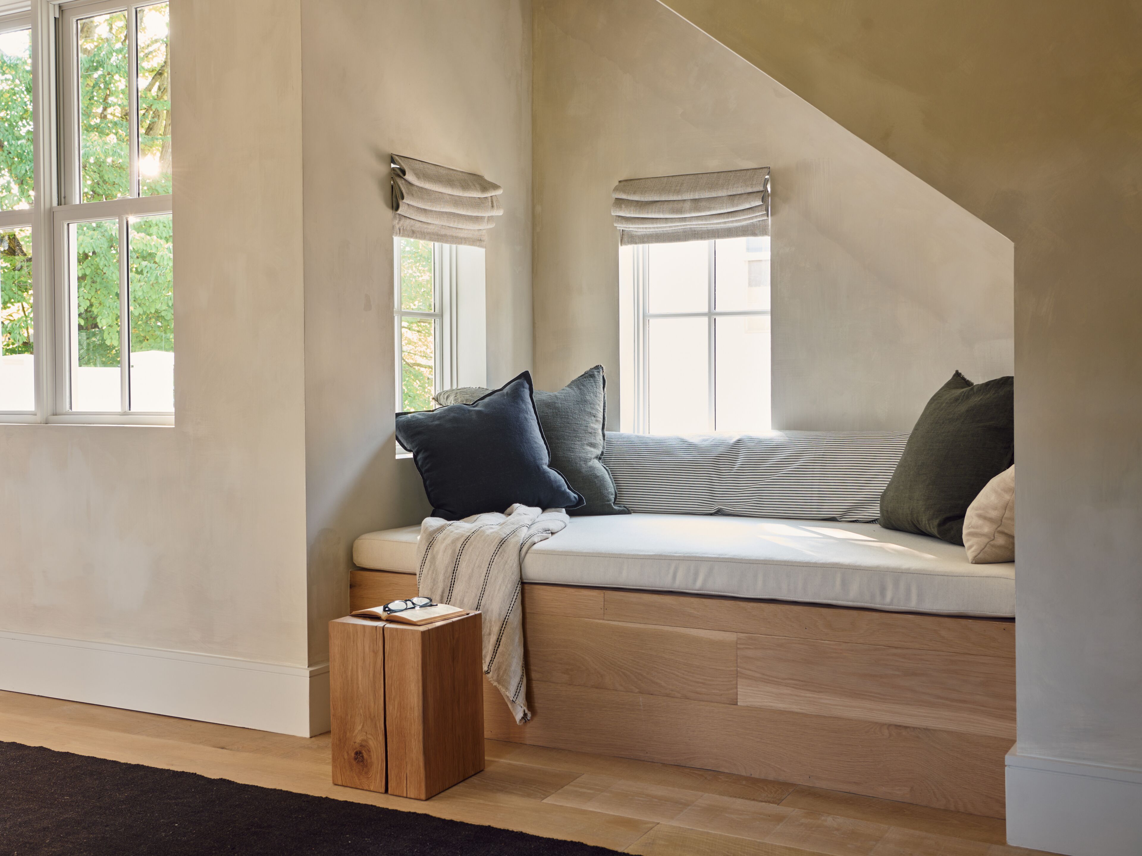 A cozy reading nook with a cushioned bench, a small wooden stool, and a window with curtains.