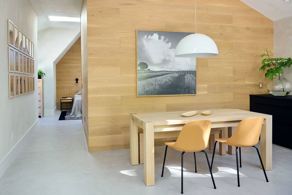 A modern dining area with a wooden table and two chairs, a pendant light, and a wall-mounted painting.
