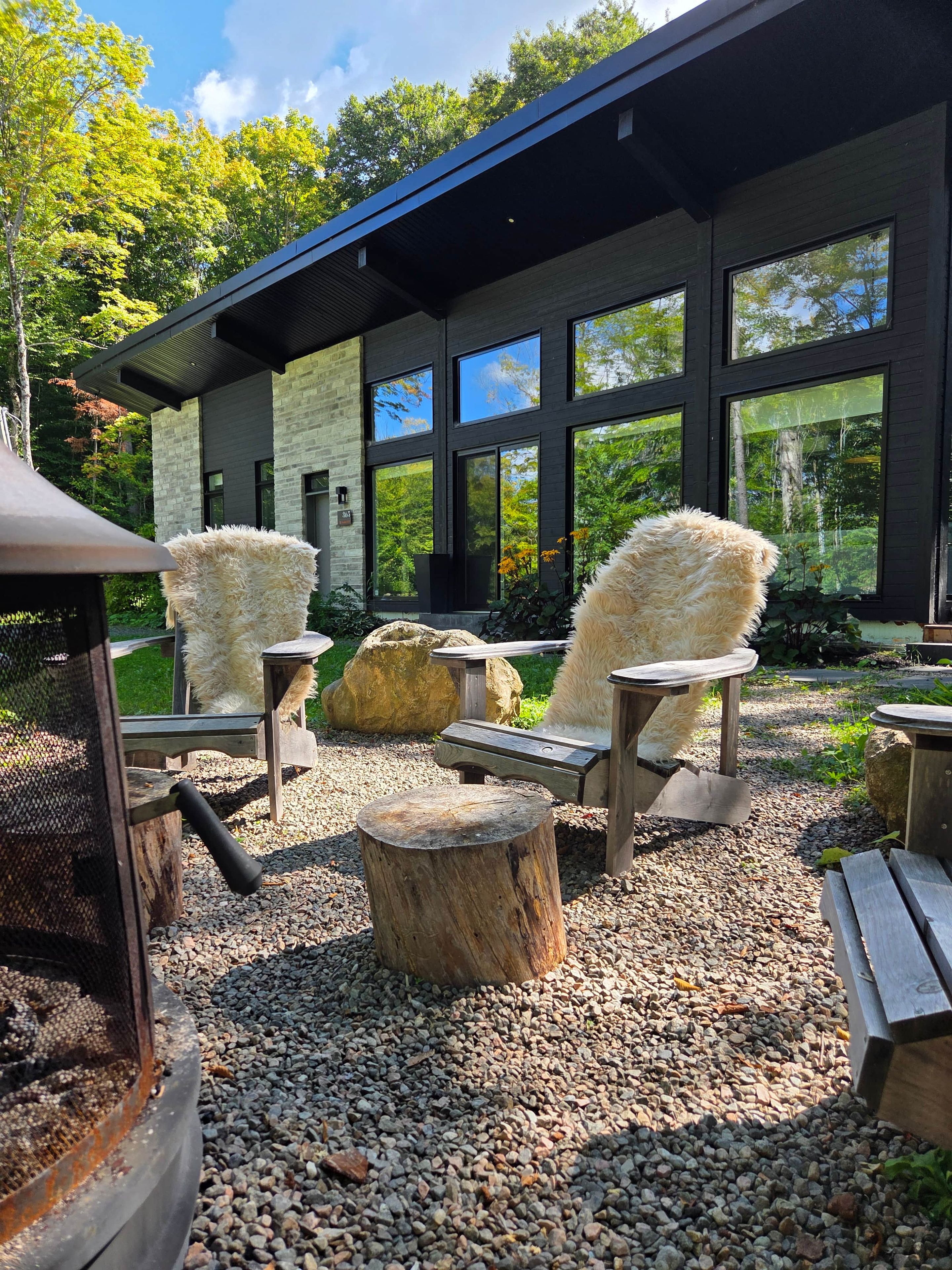 A modern outdoor seating area with chairs and a fire pit, surrounded by a gravel path and large windows overlooking a forested area.