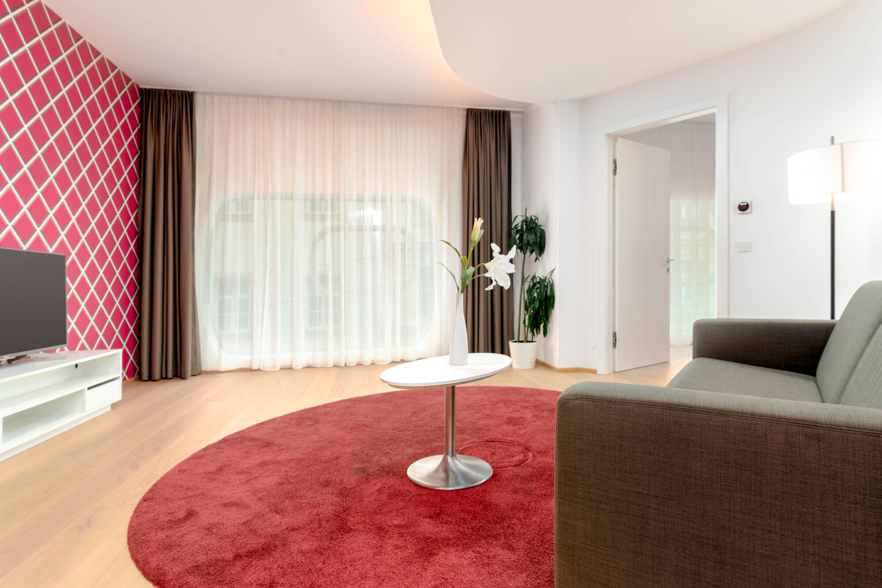 A modern living room with a red carpet, a grey sofa, a white round table, and a TV on a white stand.