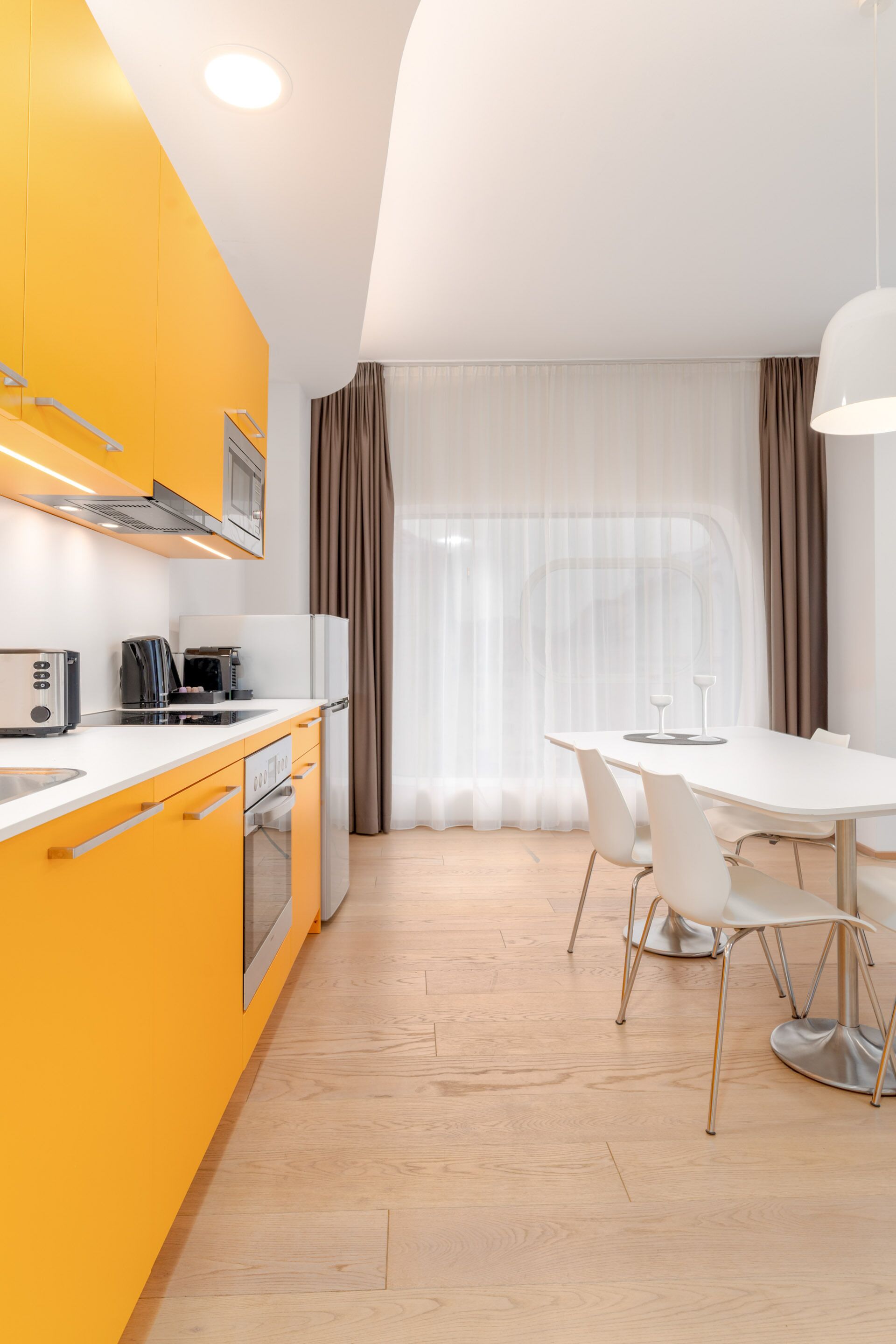 Una cocina moderna con encimera blanca, armarios amarillos y electrodomésticos de acero inoxidable. Cuenta con una zona de comedor con mesa y sillas blancas, y un ventanal con cortinas de gasa.