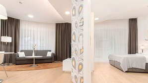 Une chambre d’hôtel moderne avec un canapé gris, une petite table ronde, un lit avec une tête de lit grise et une cloison blanche ornée d’un motif décoratif.